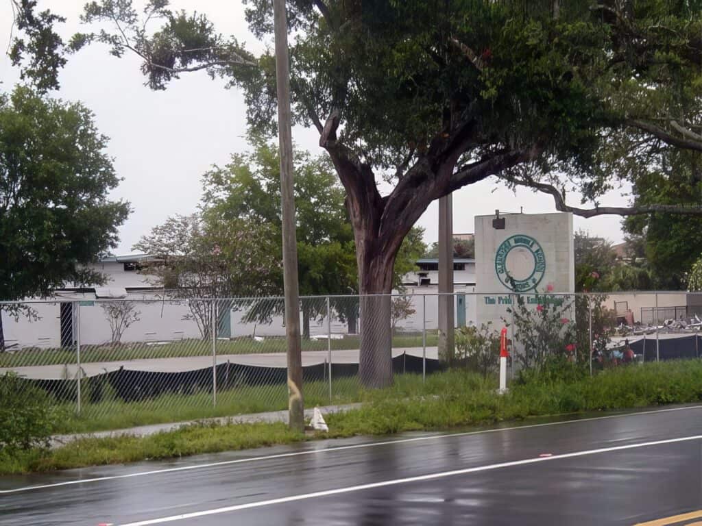 Forgotten Florida - Urbex of Glenridge Middle School 8 Tree and fence near a recreational area with a sign reading "Unfinished Man" on a cloudy day.