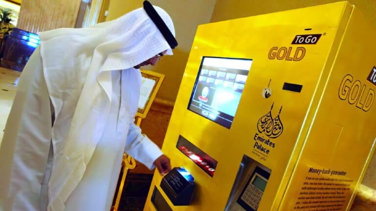 ATM machine with gold branding at Emirates Palace in Abu Dhabi, luxurious setting, man in traditional Middle Eastern attire using the ATM, gold-colored kiosk, high-end hotel environment.