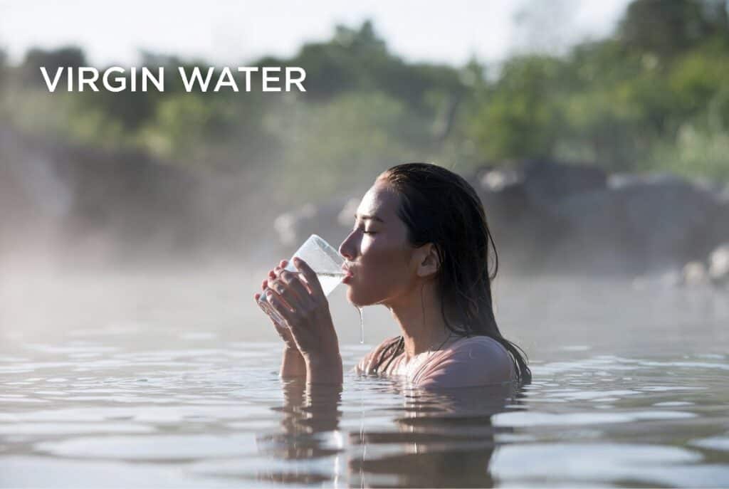 Virgin Water: The Benefits of Drinking Clean Water 2 Refreshing woman drinking from a glass of pure virgin water while immersed in natural hot spring with steam, outdoor greenery, and clear sky in the background.