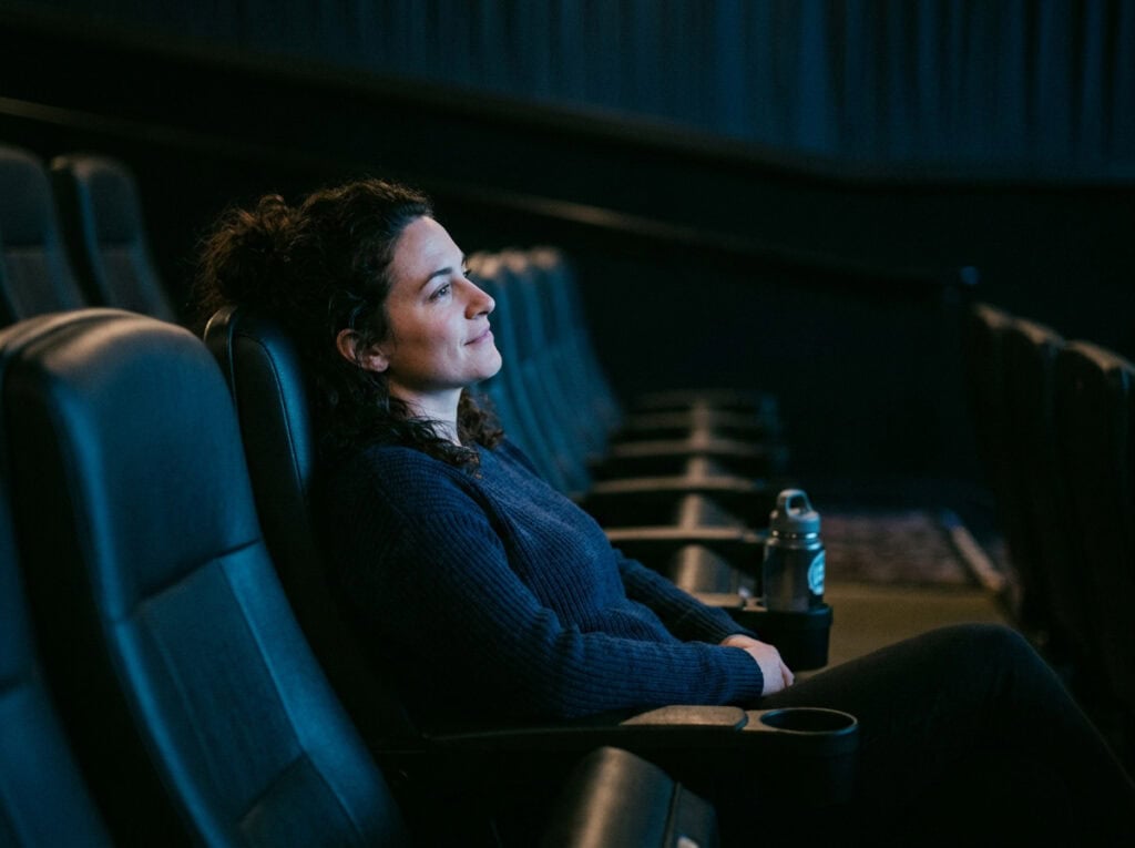Woman enjoying a solo movie night in a dark theater, sitting comfortably in a theater seat with a drink and snacks, embracing her alone time on a Friday night.