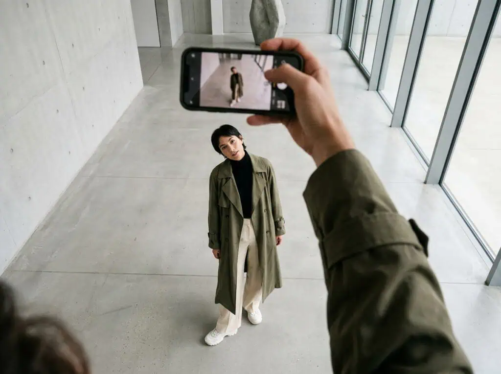 A person taking a high-quality selfie with a smartphone, capturing a fashionable individual posing confidently in a sleek, minimalist space with large windows and natural lighting.
