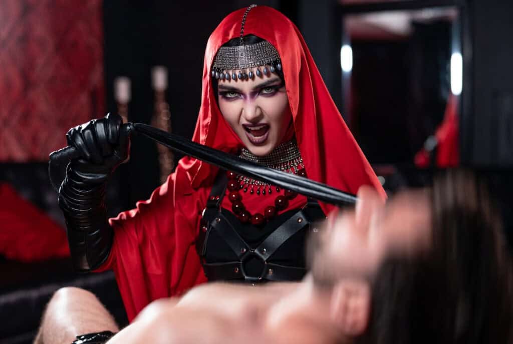 Woman dressed as a dark queen or dominatrix with a red hood, black leather outfit, and whip, portraying a powerful and intense character in a BDSM-themed setting.