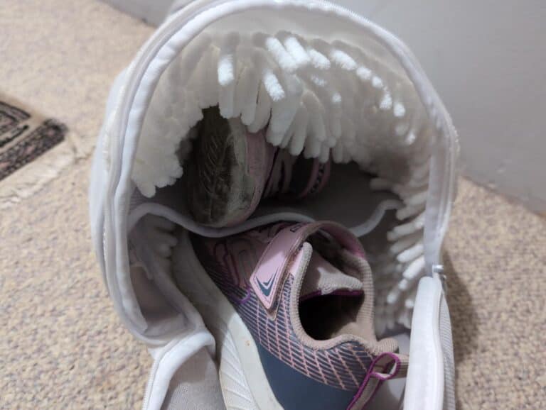 Worn athletic shoes inside a white shoe bag with soft lining, placed on beige carpet near a wall. The shoes show signs of use with dirt on the soles and scuffs, suggesting they are used for running or walking. The bag appears to be designed for convenient storage and transport of sneakers.