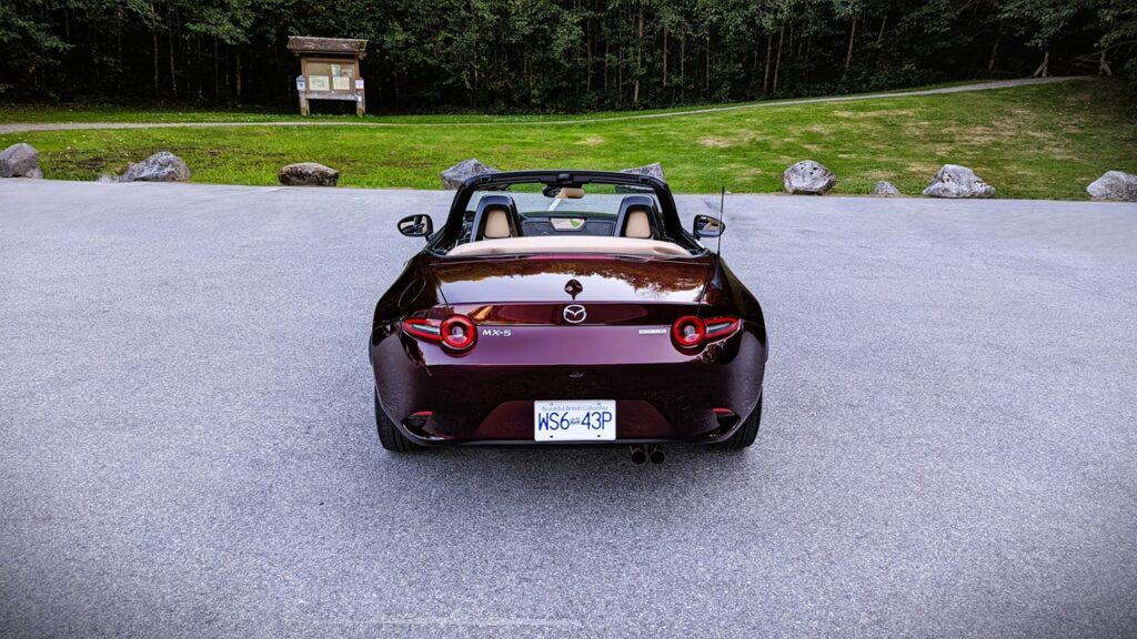 Convertible Mazda MX-5 sports car parked on a paved surface with a scenic backdrop of greenery and rocks.