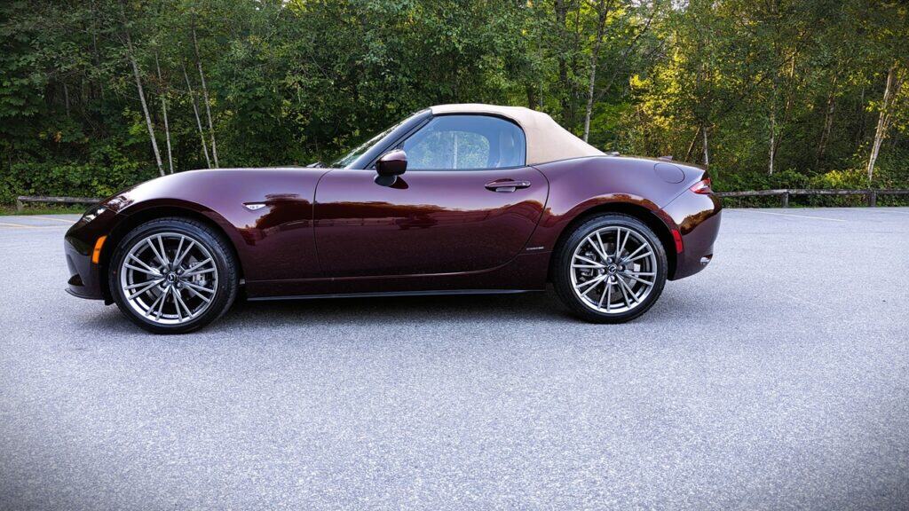 Sleek maroon convertible sports car parked on a scenic road with lush green trees in the background.