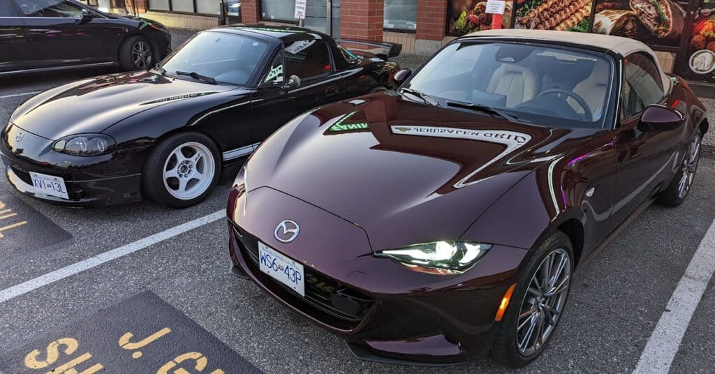 Mazda MX-5 Miata sports car parked in a lot with a black classic car nearby, modern urban setting, sleek design, and shiny exterior.