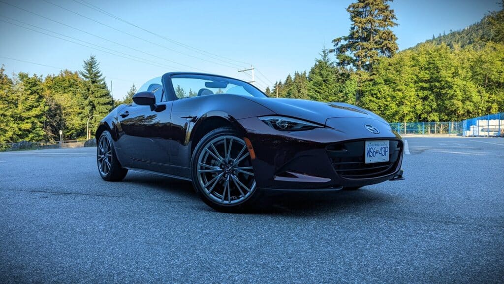 Convertible sports car parked on an empty asphalt lot with green trees and blue sky in the background.