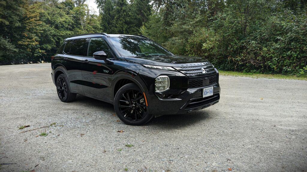 2025 Mitsubishi Outlander PHEV Noir Review 6 Black SUV parked on a gravel road surrounded by green trees, modern design, sleek exterior, urban outdoor setting, showcasing the latest vehicle model from Unfinished Man.