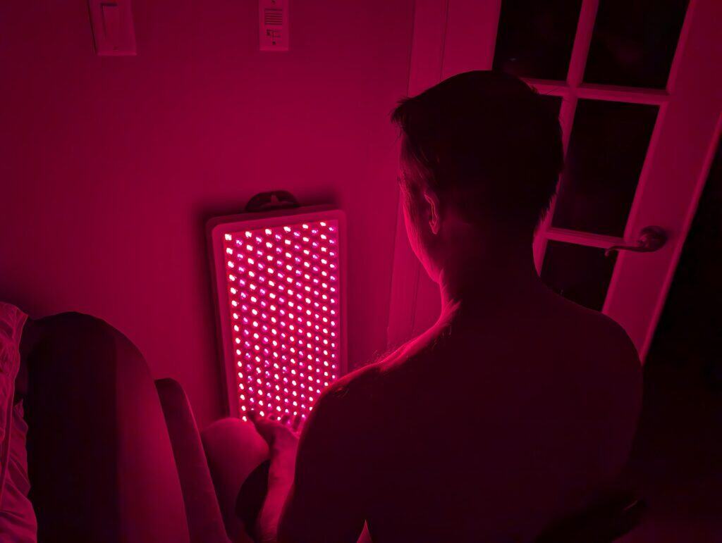 BestQool Red Light Therapy Pro300 Review: 9 Bright Benefits 2 Red LED light therapy device emitting pink glow, used for skin treatment or wellness routines, visible in a cozy bedroom setting with a man facing away from the camera.