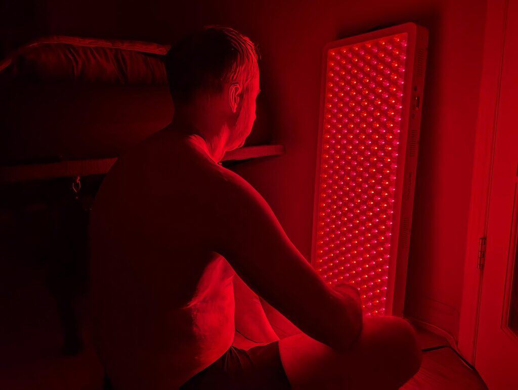 BestQool Red Light Therapy Pro300 Review: 9 Bright Benefits 8 Red LED therapy device emitting red light for skin treatment or wellness benefits, with a man sitting nearby in a dark room illuminated by the device.