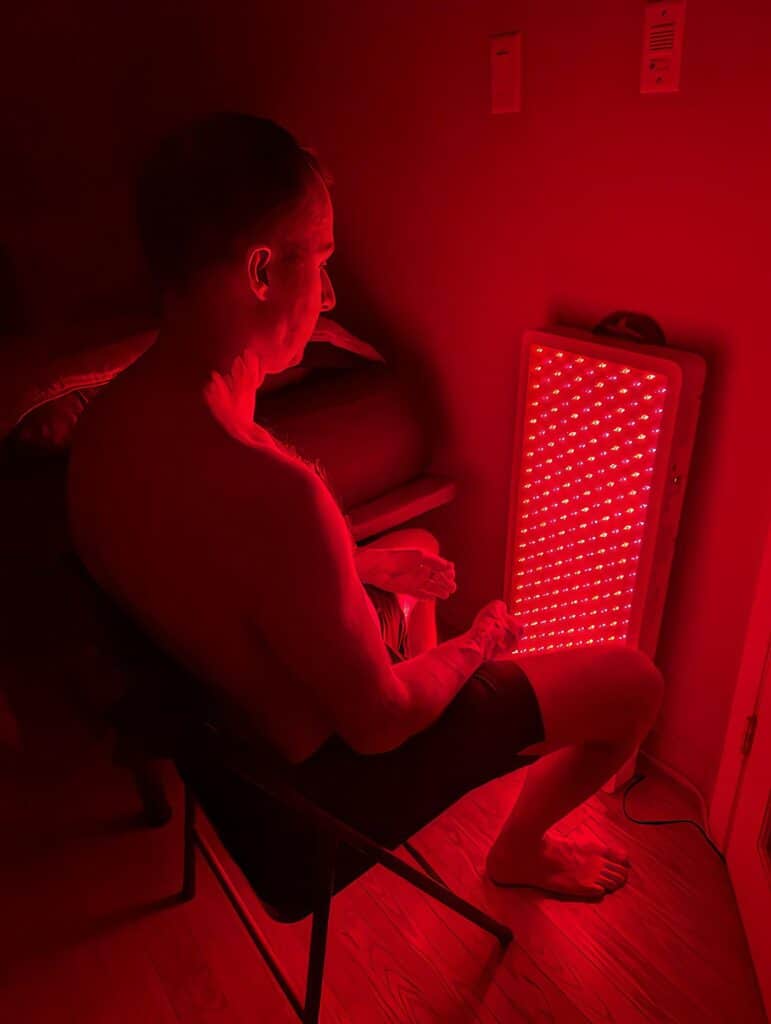 BestQool Red Light Therapy Pro300 Review: 9 Bright Benefits 13 Red light therapy device used by a man sitting on a chair in a dimly lit room for relaxation or skin health benefits.