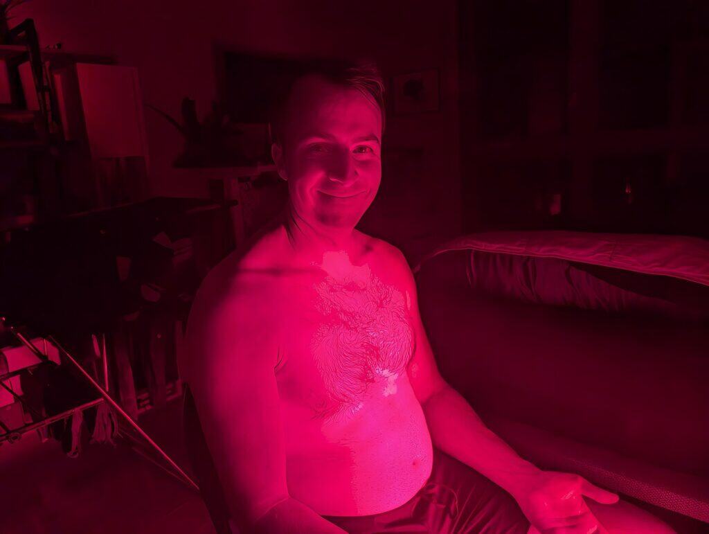 BestQool Red Light Therapy Pro300 Review: 9 Bright Benefits 14 Brightly lit man with chest tattoo smiling underwater in a pink hue, sitting in a room with shelves and a bed, embracing a relaxed and confident vibe.