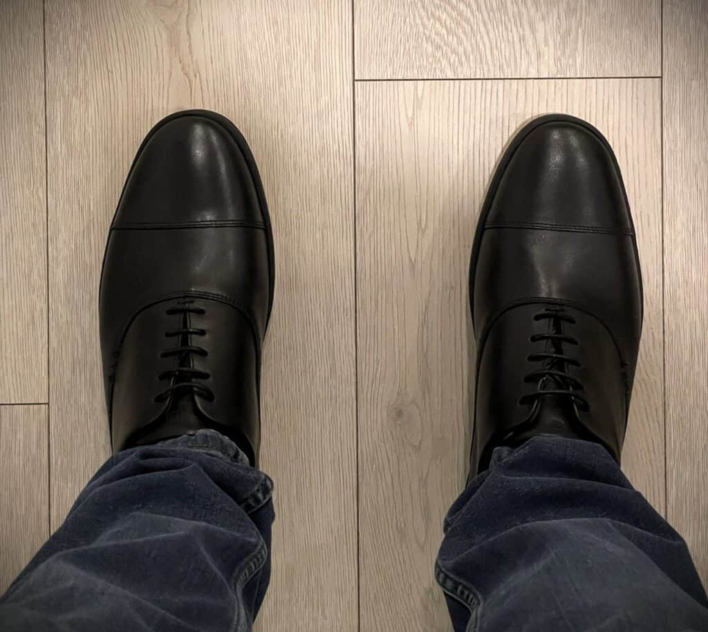 Deming Recovery 'The Westsider' Shoes Review 7 Shiny black leather dress shoes on a person's feet, viewed from above, standing on light wood flooring, stylish men's fashion footwear for formal or professional settings.