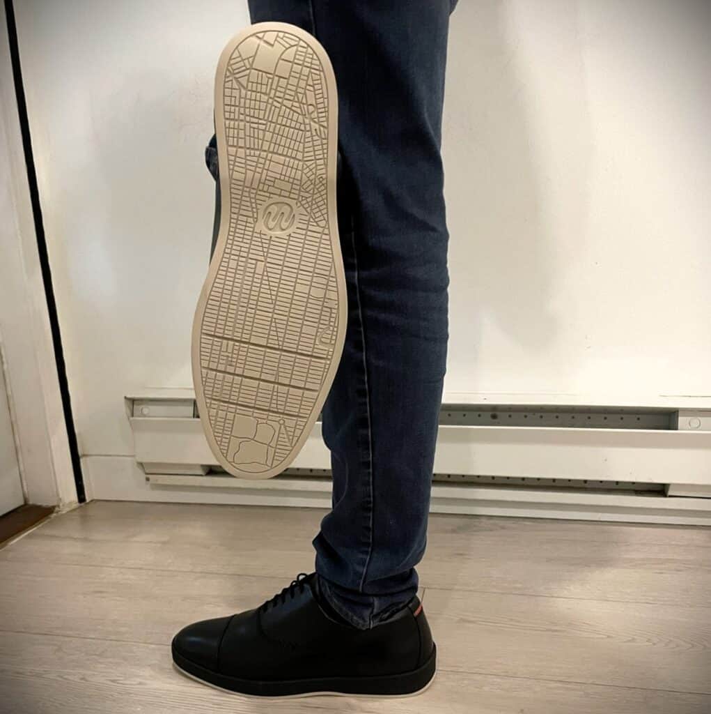 Deming Recovery 'The Westsider' Shoes Review 4 Stylish black sneakers on a person's feet, one lifted to show the sole with a grid pattern and logo, paired with dark jeans, standing on light-colored wooden flooring in an indoor setting.