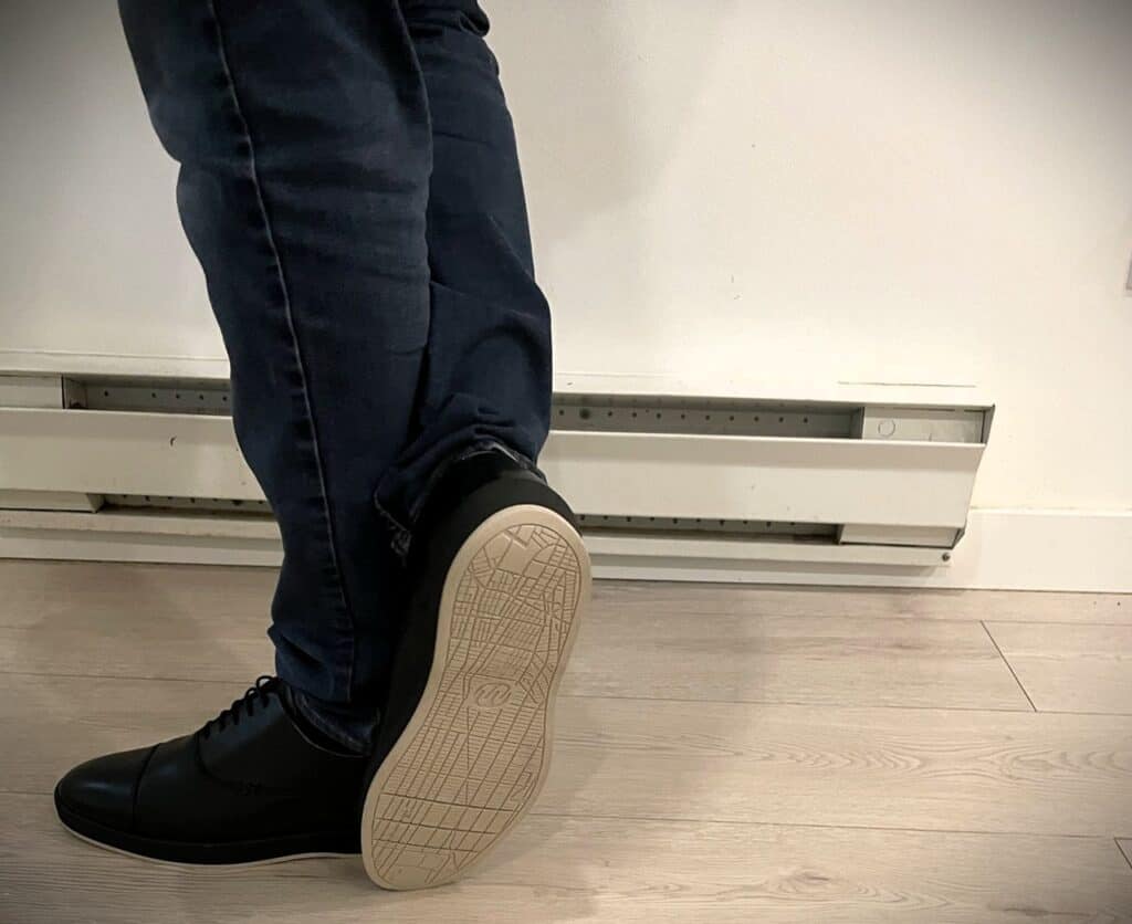 Deming Recovery 'The Westsider' Shoes Review 2 Stylish black sneakers worn with dark jeans, standing on a light wood floor near a baseboard heater. Modern men's footwear and casual fashion in an indoor setting.