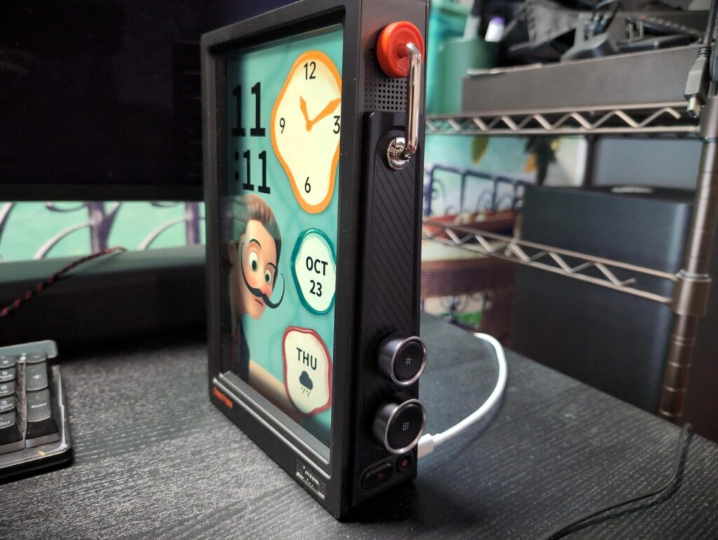 Divoom Times Frame Review 7 Digital alarm clock with a cartoon face display, showing the time, date, and day of the week on a black desk surface, with a keyboard and shelf in the background.