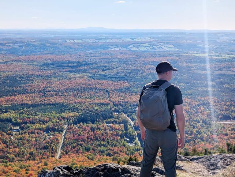 Vast mountain landscape overlooking colorful autumn forest with man in black shirt, gray pants, and backpack enjoying scenic view from peak.