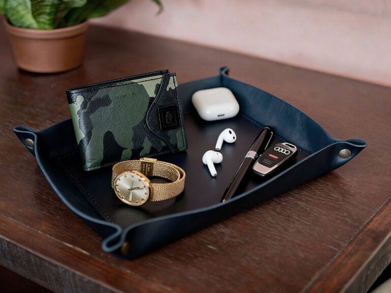 Camo wallet, gold watch, wireless earbuds, AirPods case, and car key on leather tray on wooden desk.
