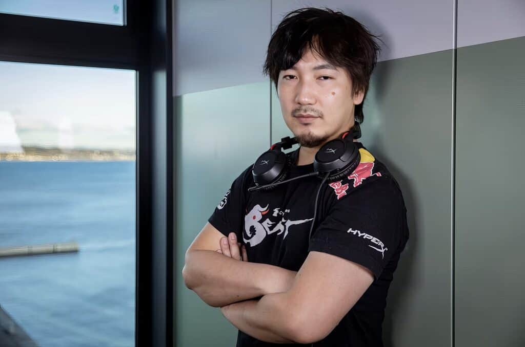 Daigo Umehara: The Legendary Beast's Complete Fighting Game Journey 6 Headphones around neck of confident man with casual Black T-shirt, standing near window with water view, modern indoor setting, casual style, contemporary lifestyle, technology.