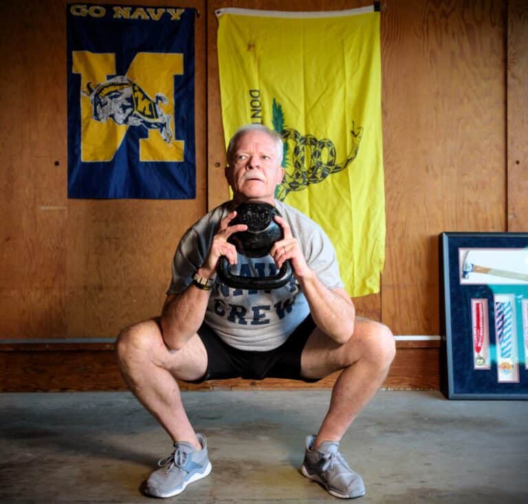 Strong man performing an indoor squat exercise with a kettlebell, surrounded by military flags, exhibiting fitness and strength training.