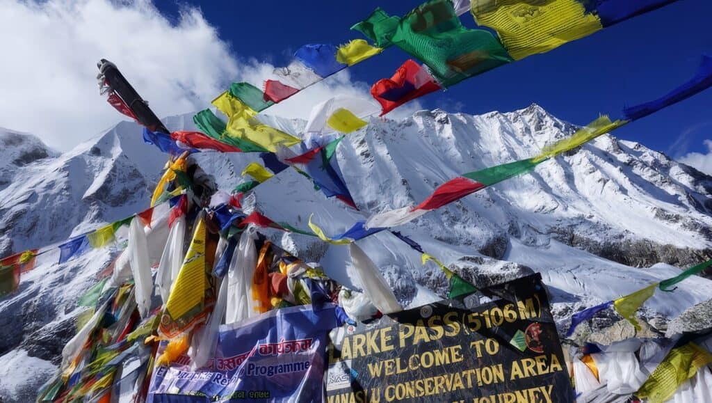 How Difficult Is Manaslu Trek? Challenges & Tips 7 Colorful prayer flags fluttering against a snow-covered mountain peaks in Nepal, near Larke Pass at 5106 meters, welcoming travelers to the conservation area on a clear, sunny day.