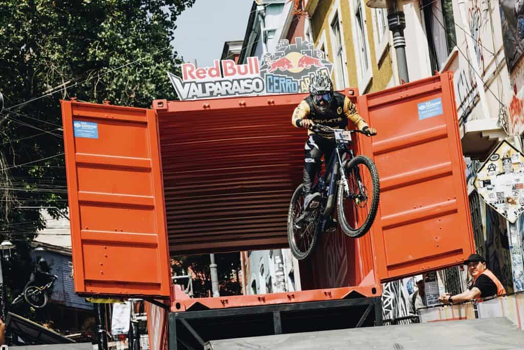 A man in his mid-30s rides his bike down a steep, worn street in Valparaíso. Extreme mountain biking jump off a large container at Red Bull Valparaíso Cerro Abajo urban downhill event in Valparaíso, Chile.