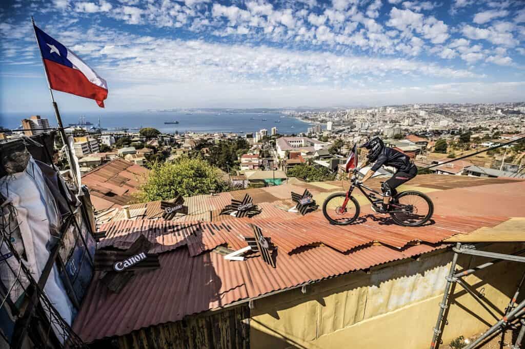 A young man casually rides his bike down a colorful mural-lined stairway in Valparaíso. Colorful cityscape of Santiago, Chile, with a mountain biking enthusiast riding on a rooftop with a panoramic view of the city and ocean in the background.