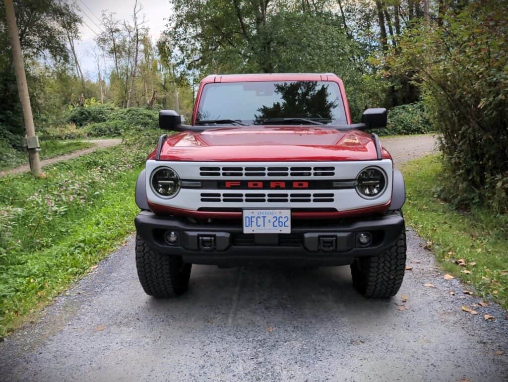 A red Ford Bronco SUV parked on a gravel trail surrounded by lush greenery and trees, showcasing rugged off-road capability and adventure-ready design.