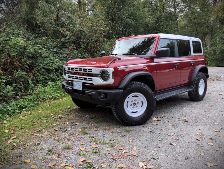 A classic Ford Bronco SUV parked on a gravel trail surrounded by lush green trees and foliage, showcasing its vintage design, rugged tires, and red and white exterior, ideal for outdoor adventures and car enthusiasts.