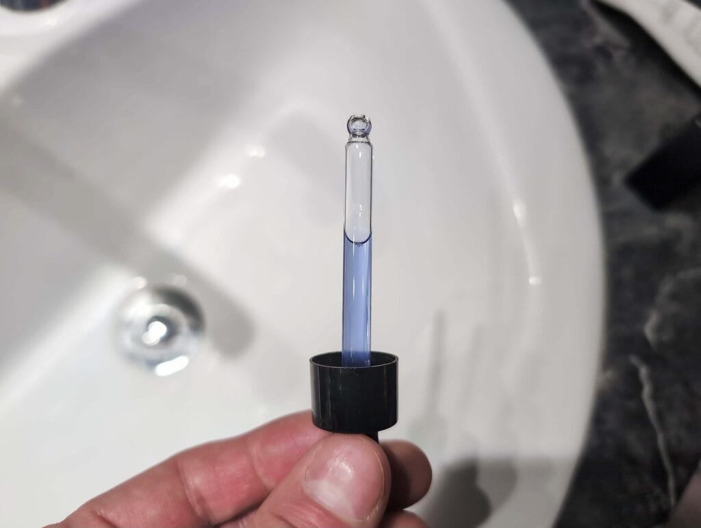 Are Copper Peptides Worth It For Skin And Hair? 2 A clear glass test tube with a blue liquid, held by a person's hand above a bathroom sink with a silver drain, showcasing scientific experimentation or laboratory work.