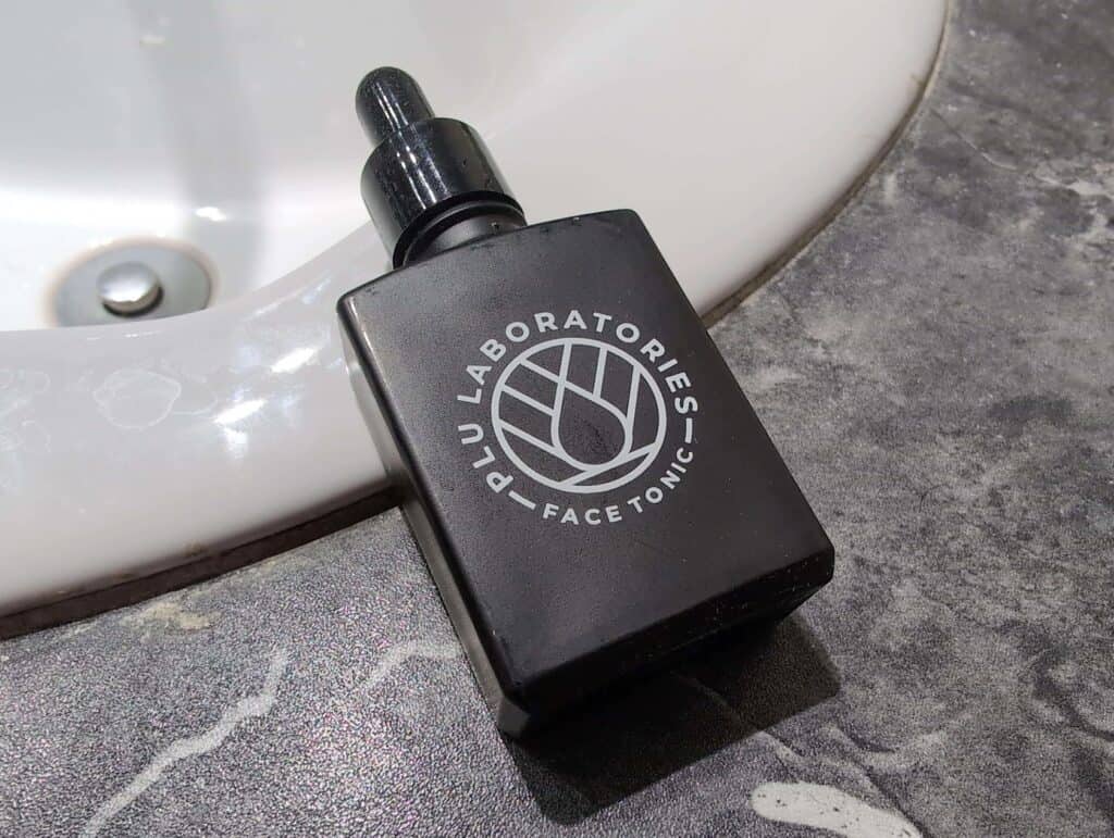 Are Copper Peptides Worth It For Skin And Hair? 3 A black bottle of Clinique facial toner with the label "PLU LABORATORIES FACE TONIC" resting on a gray textured bathroom countertop next to a white sink.