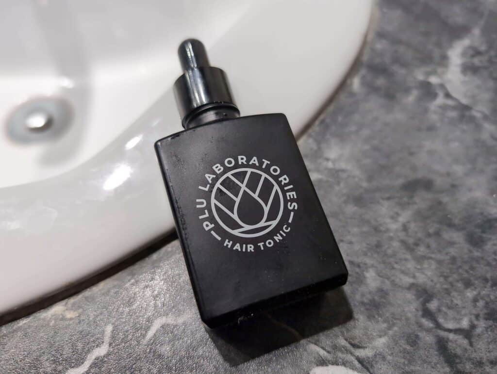 Are Copper Peptides Worth It For Skin And Hair? 4 Black hair tonic bottle with logo on a marble countertop in a bathroom.