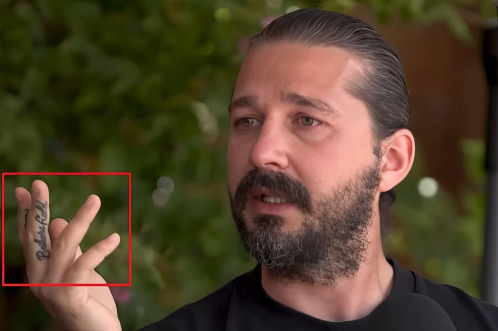 Portrait of a mature man with a beard and slicked-back hair, gesturing with his right hand that has the word "BLURRED" tattooed on his fingers. The background features blurred greenery, suggesting an outdoor setting.