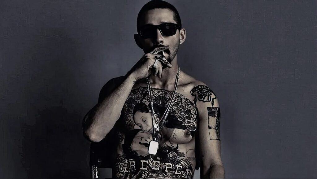 A tattooed man in sunglasses, adorned with necklaces and rings, striking a confident pose. His tattoos and fashion style reflect masculinity and modern urban culture.