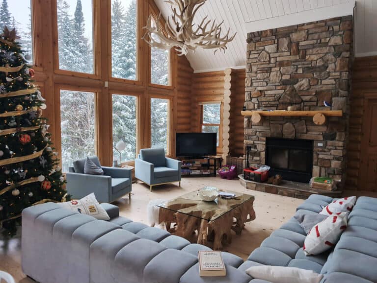 Bright, rustic living room featuring large windows with snowy forest views, stone fireplace, and cozy furniture. Perfect for holiday celebrations and winter comfort.