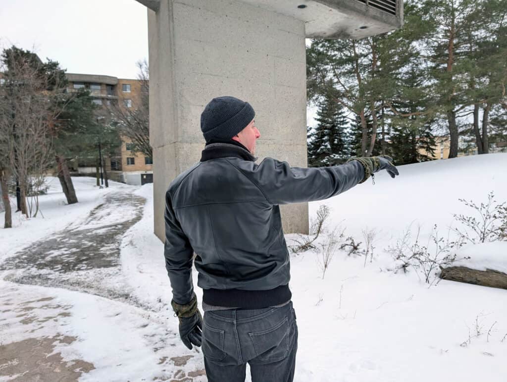 Lusso Leather Black Bomber Style Ribbed Jacket Review 13 A man dressed in winter clothing, including a black beanie, gloves, and a leather jacket, standing near a concrete structure in a snowy outdoor setting, pointing towards the snow and trees.