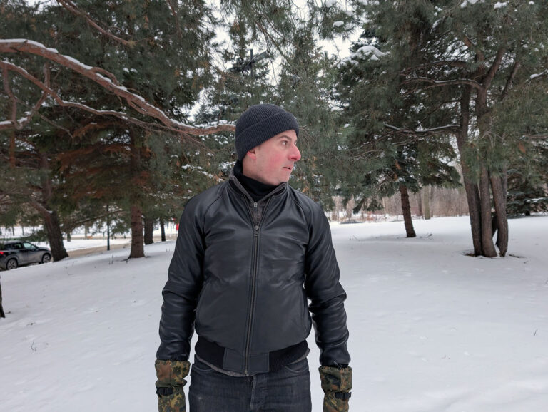 A man dressed warmly in a black leather jacket, black beanie, and camouflage gloves stands in a snowy landscape with pine trees, capturing a stylish winter look perfect for outdoor winter fashion inspiration.