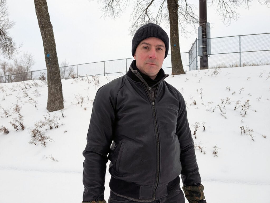 Lusso Leather Black Bomber Style Ribbed Jacket Review 2 A man dressed in black winter jacket and beanie stands outdoors in snowy landscape, surrounded by bare trees and overcast sky, embodying style and resilience for men's winter fashion.