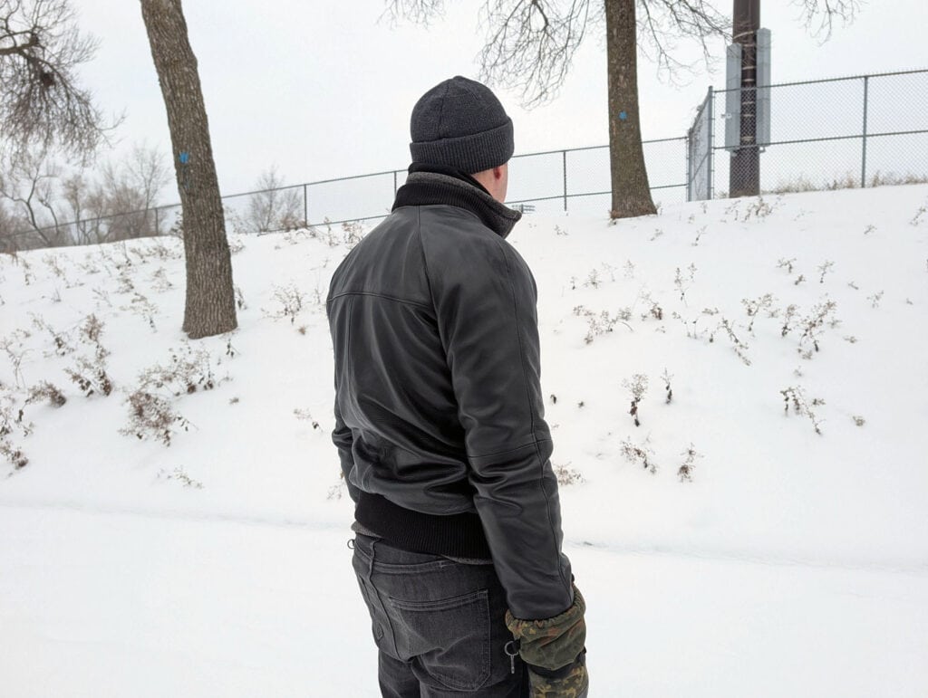 Lusso Leather Black Bomber Style Ribbed Jacket Review 3 Man wearing black leather jacket and beanie in snowy outdoor park scene, winter landscape with trees, snow, and a fence, cold weather fashion and winter apparel.