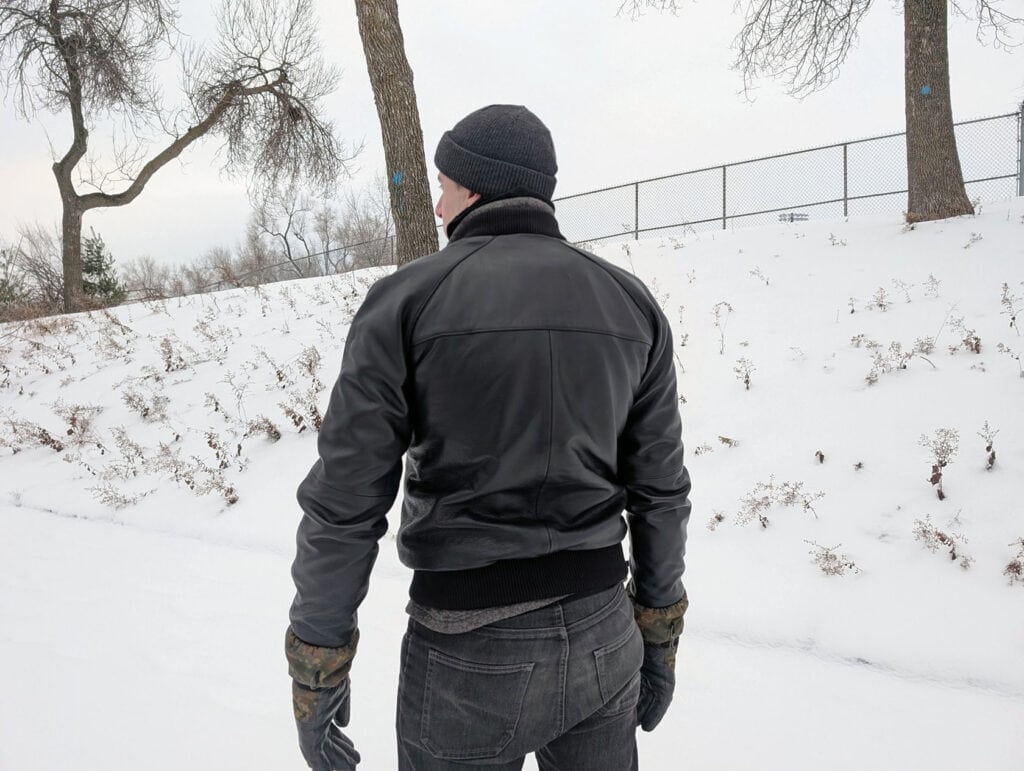 Lusso Leather Black Bomber Style Ribbed Jacket Review 4 A man walks in a snowy outdoor scene wearing a black leather jacket, black beanie, and winter gloves, highlighting stylish winter apparel for men.