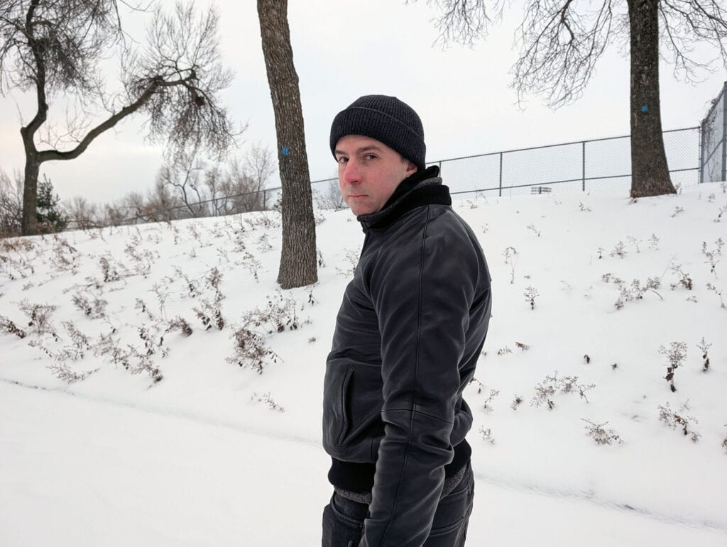 Lusso Leather Black Bomber Style Ribbed Jacket Review 5 Male in black winter jacket and beanie, standing in snowy landscape with trees, cold weather clothing, winter outdoor apparel for men.