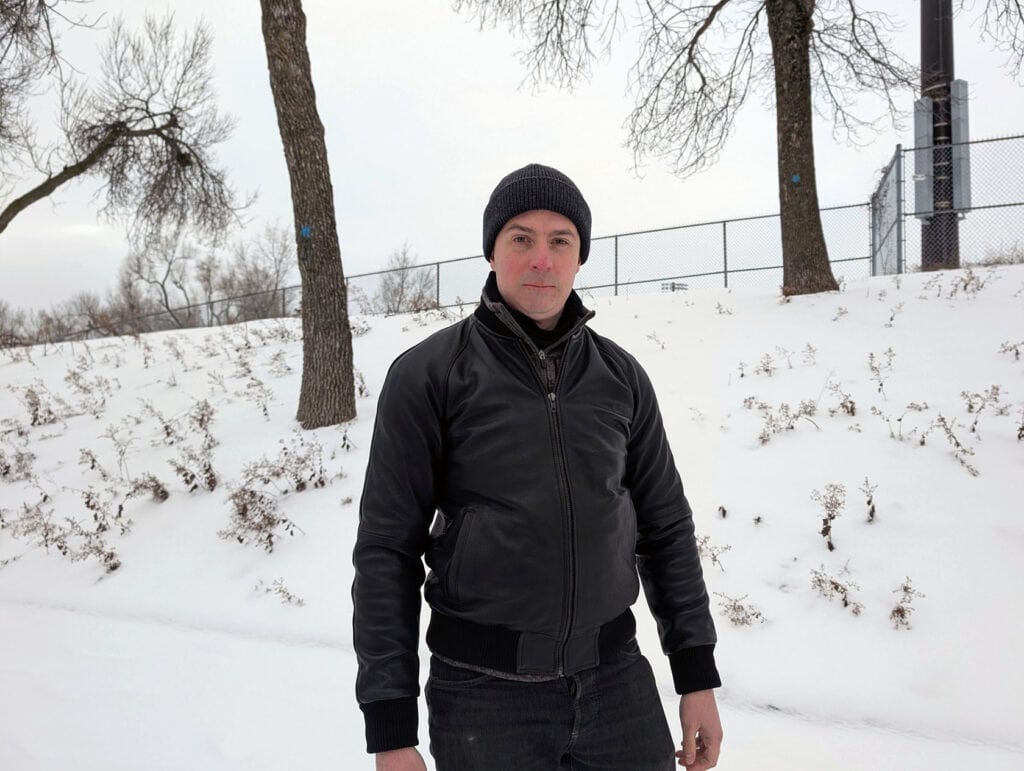 Lusso Leather Black Bomber Style Ribbed Jacket Review 7 A man dressed in a black leather jacket and beanie hat standing in a winter outdoor setting with snow-covered ground, barren trees, and a fenced area, capturing a stylish and rugged winter look for men's fashion and lifestyle content.