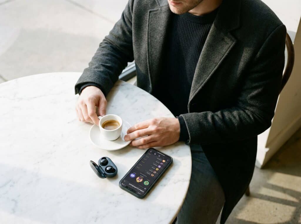 Close-up of a man in a dark blazer drinking espresso while using wireless earbuds and a mobile app on his phone at a stylish cafe.