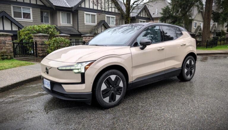 An image of the 2026 Volvo EX30 Twin Motor, showcasing its sleek design and modern features, parked on a wet street with residential houses in the background.