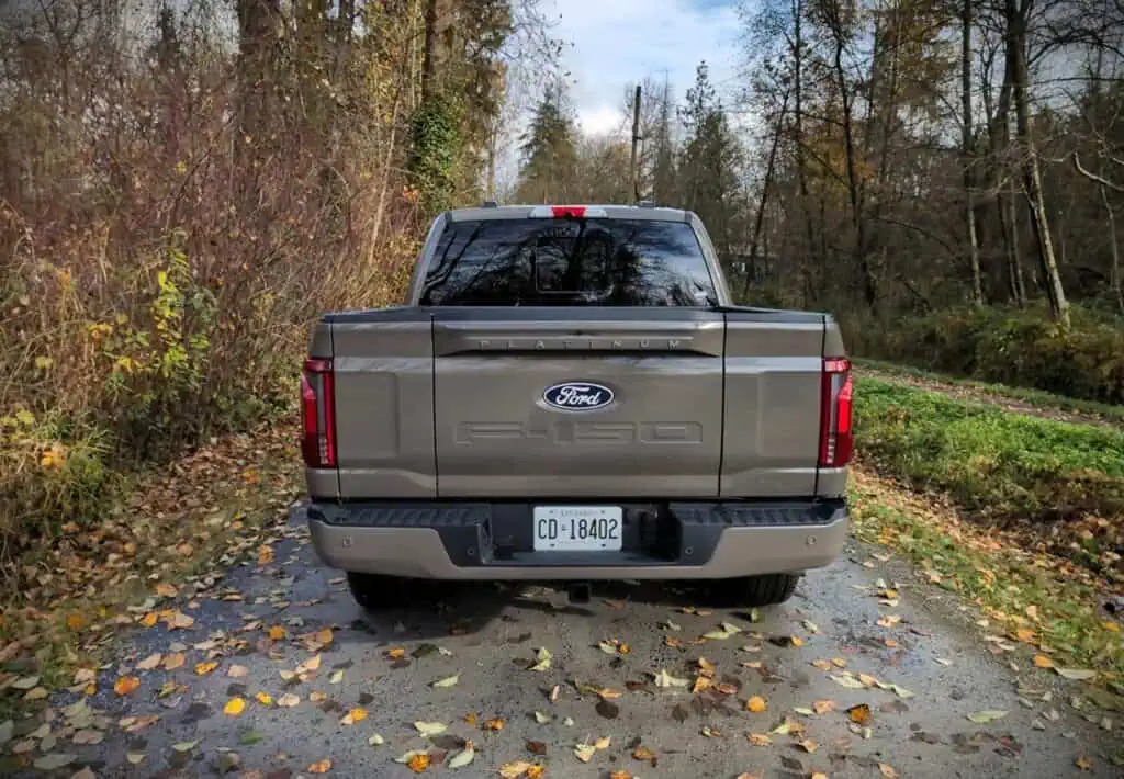 An image of the rear view of a 2026 Ford F150 SuperCrew Platinum Hybrid pickup truck parked on a leaf-covered road surrounded by trees.