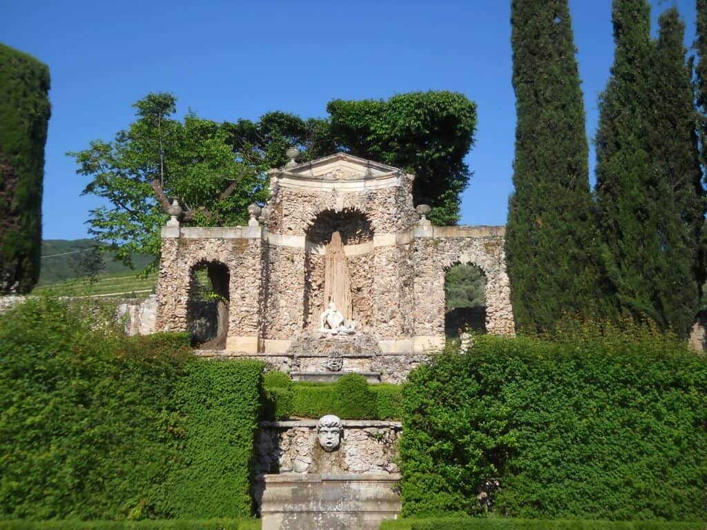 Ruins of an ancient Italian garden with stone structures, lush greenery, and tall cypress trees, showcasing Italy's historic landscape and garden architecture.