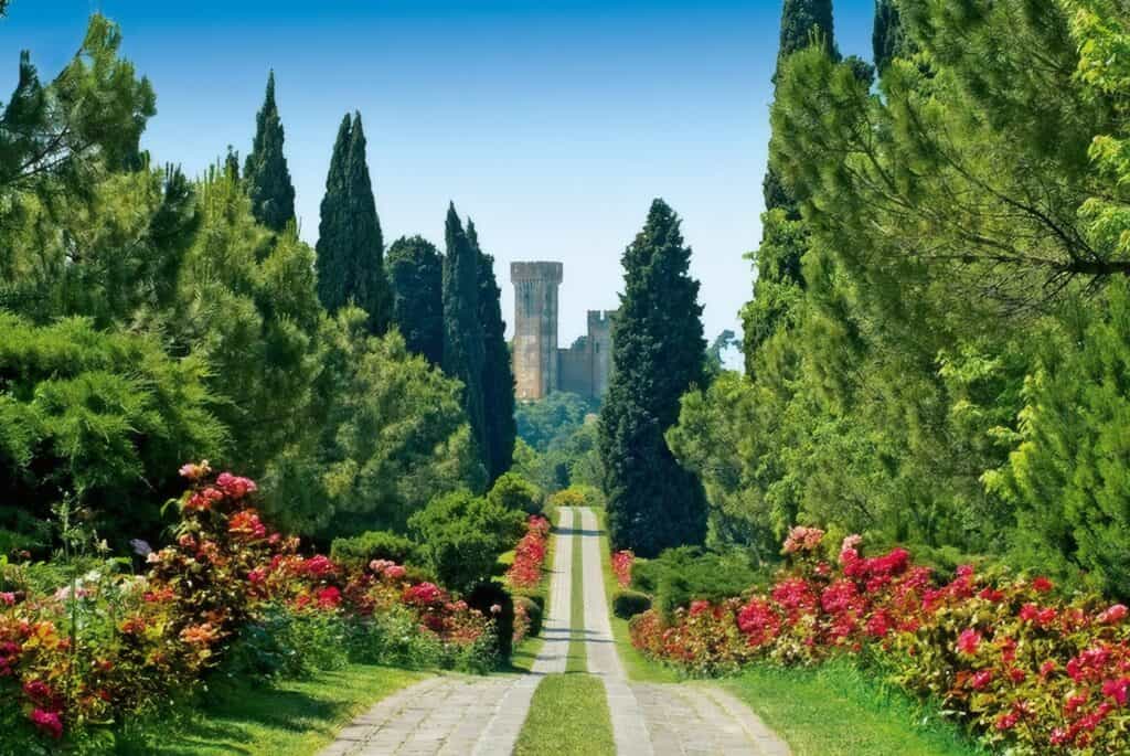Beautiful historic garden in Italy with lush greenery, colorful flowers, and tall cypress trees lining a stone pathway leading to distant castle ruins.