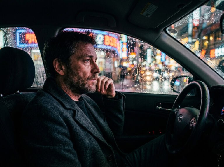 A man sitting alone in a car during rainy night, reflecting on the emotional fallout of a DUI, with city lights blurred outside, highlighting feelings of regret and contemplation.