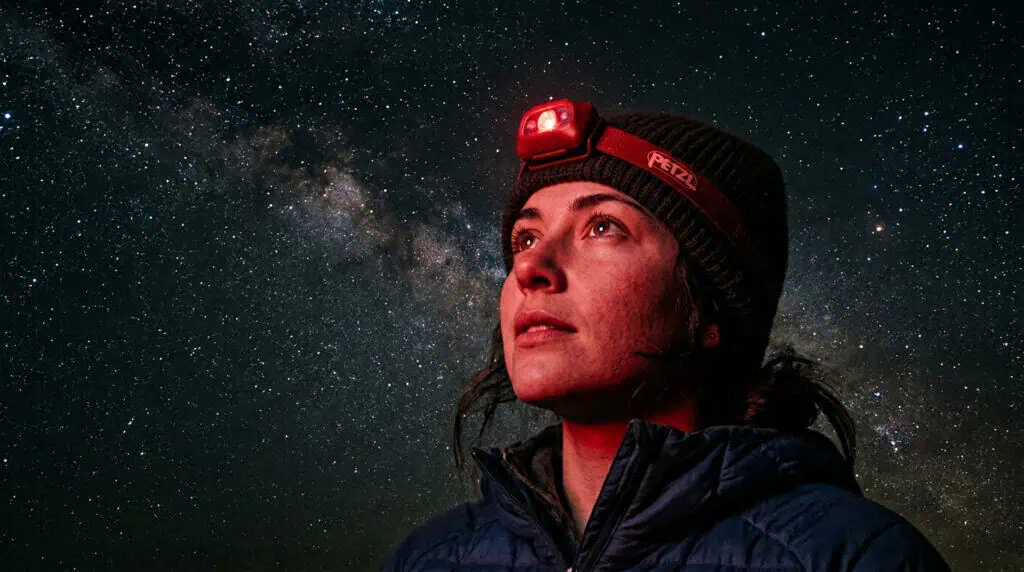 Woman wearing a Petzl headlamp under a starry night sky, ideal for camping lighting setups that avoid blinding lumens. Perfect for outdoor adventures and nighttime activities.