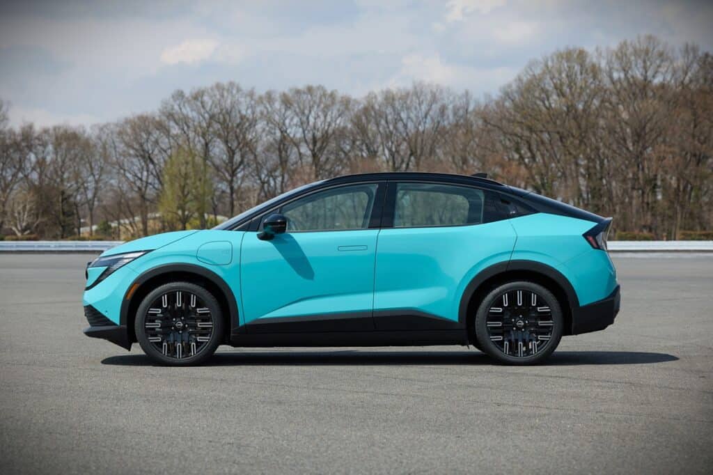 An image of a 2026 Nissan Leaf SV+ electric car parked on an open road with a backdrop of trees and a partly cloudy sky, highlighting its modern design and eco-friendly features.